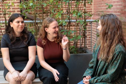 Three young women seated outside talking in front of a brick building