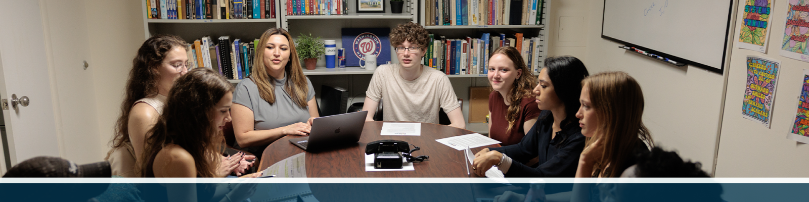 A group of students and a professor seated at a shared table with shelves of books behind them